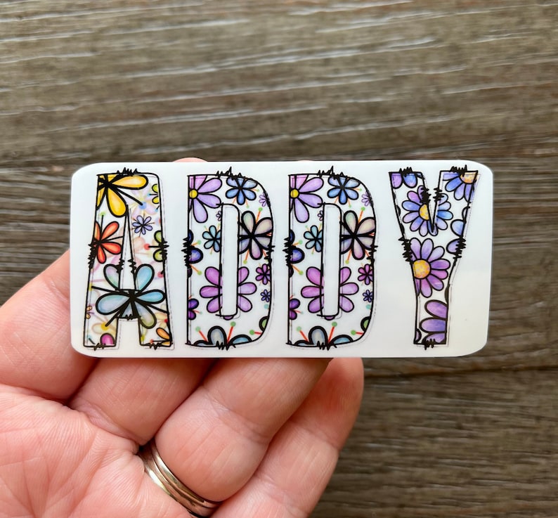 Pode incluir: Um adesivo branco com o nome "ADDY" escrito em letras com contorno preto. As letras s&atilde;o preenchidas com um padr&atilde;o floral colorido de flores roxas, azuis, amarelas e rosas.