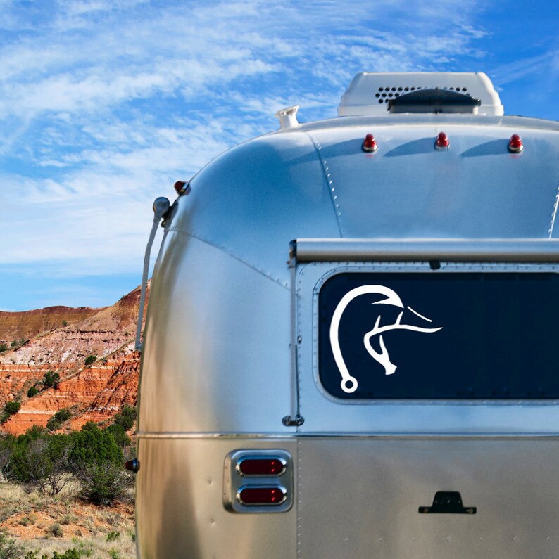 K&ouml;nnte beinhalten: Ein wei&szlig;er Airstream-Wohnwagen mit einem wei&szlig;en Aufkleber in Form eines Entenkopfes auf dem Heckfenster. Der Wohnwagen steht vor einer roten Felsenschlucht.