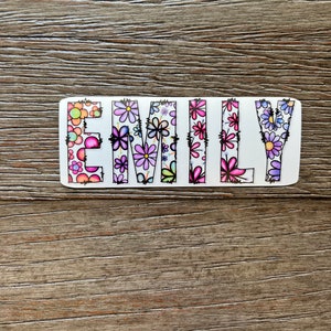 Pode incluir: Uma placa de madeira branca com o nome "EMILY" escrito em letras coloridas. As letras s&atilde;o decoradas com um padr&atilde;o floral em tons de rosa, roxo e verde.