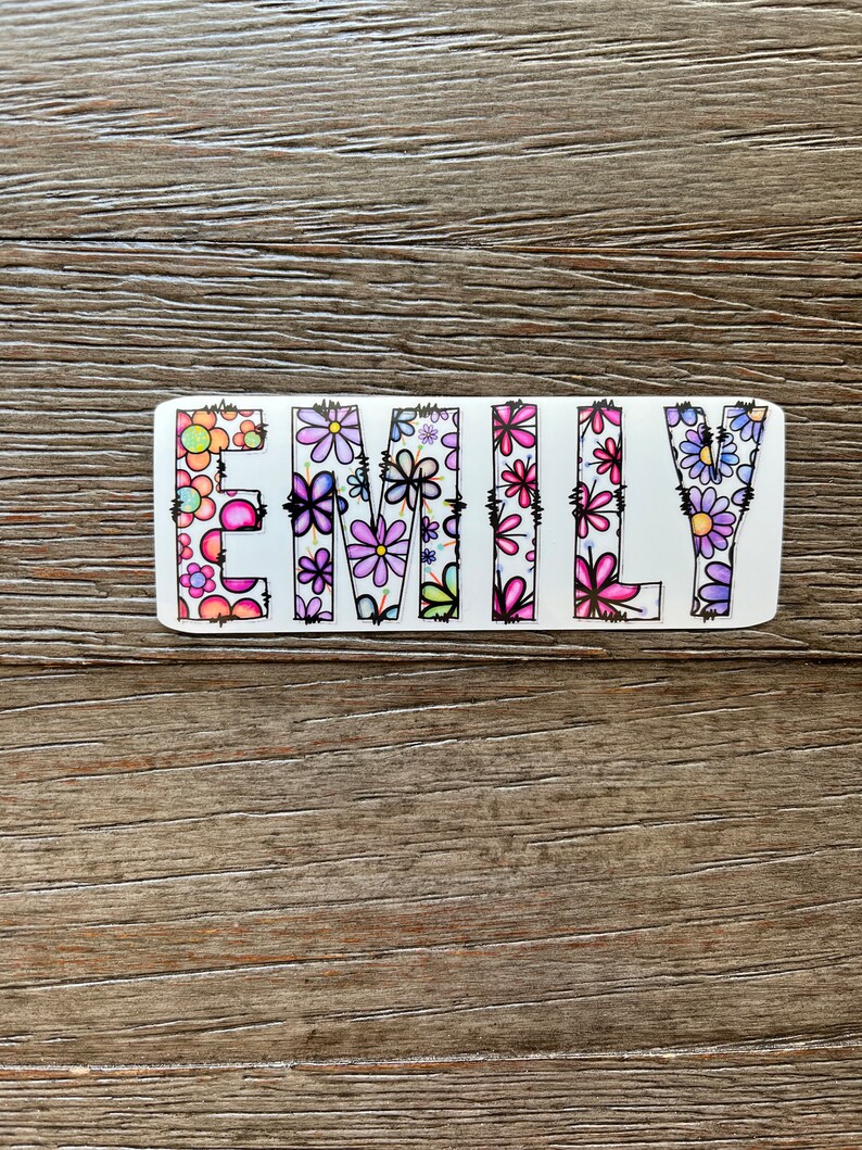 Pode incluir: Uma placa de madeira branca com o nome "EMILY" escrito em letras coloridas. As letras s&atilde;o decoradas com um padr&atilde;o floral em tons de rosa, roxo e verde.