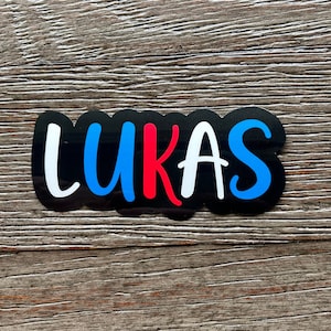 May include: A black sticker with the name "LUKAS" written in white, blue, and red letters.