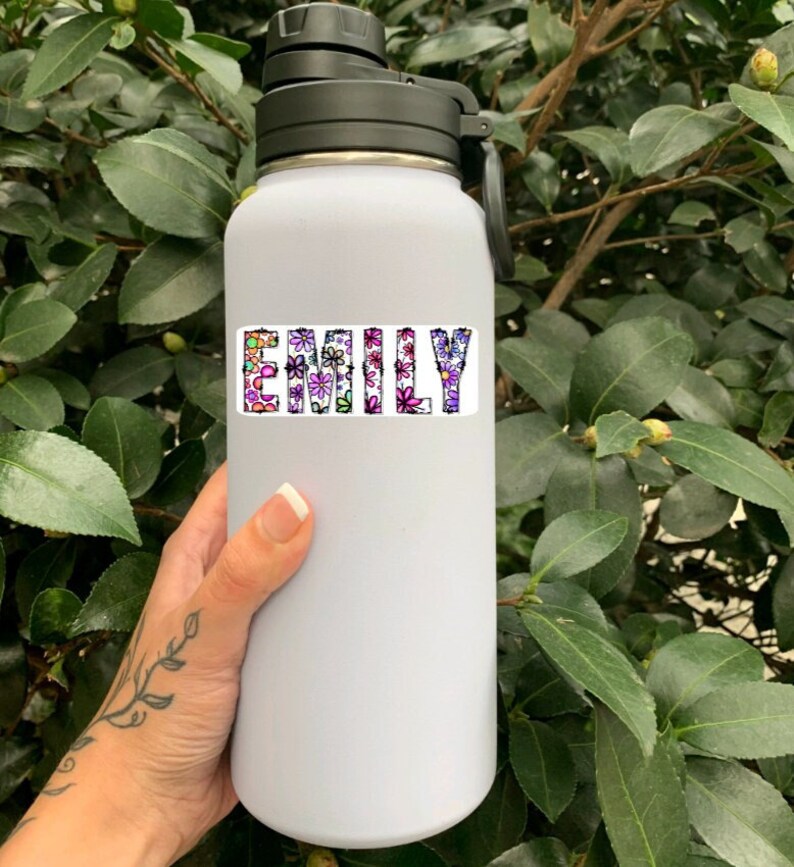 Pode incluir: Garrafa de &aacute;gua branca com tampa e al&ccedil;a pretas. O nome "Emily" est&aacute; escrito na garrafa com flores coloridas.