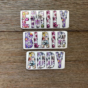 Pode incluir: Tr&ecirc;s adesivos retangulares brancos com os nomes "Emily", "Blair" e "Addy" escritos em letras pretas delineadas. As letras s&atilde;o preenchidas com um padr&atilde;o floral colorido.