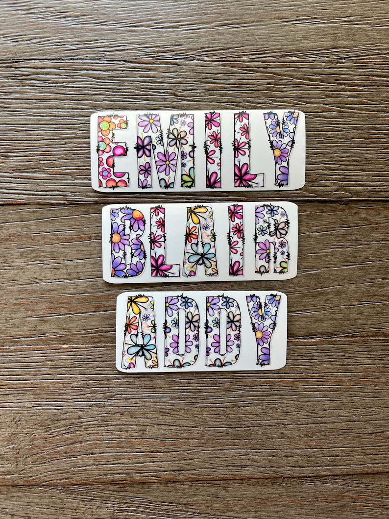 Pode incluir: Tr&ecirc;s adesivos retangulares brancos com os nomes "Emily", "Blair" e "Addy" escritos em letras pretas delineadas. As letras s&atilde;o preenchidas com um padr&atilde;o floral colorido.