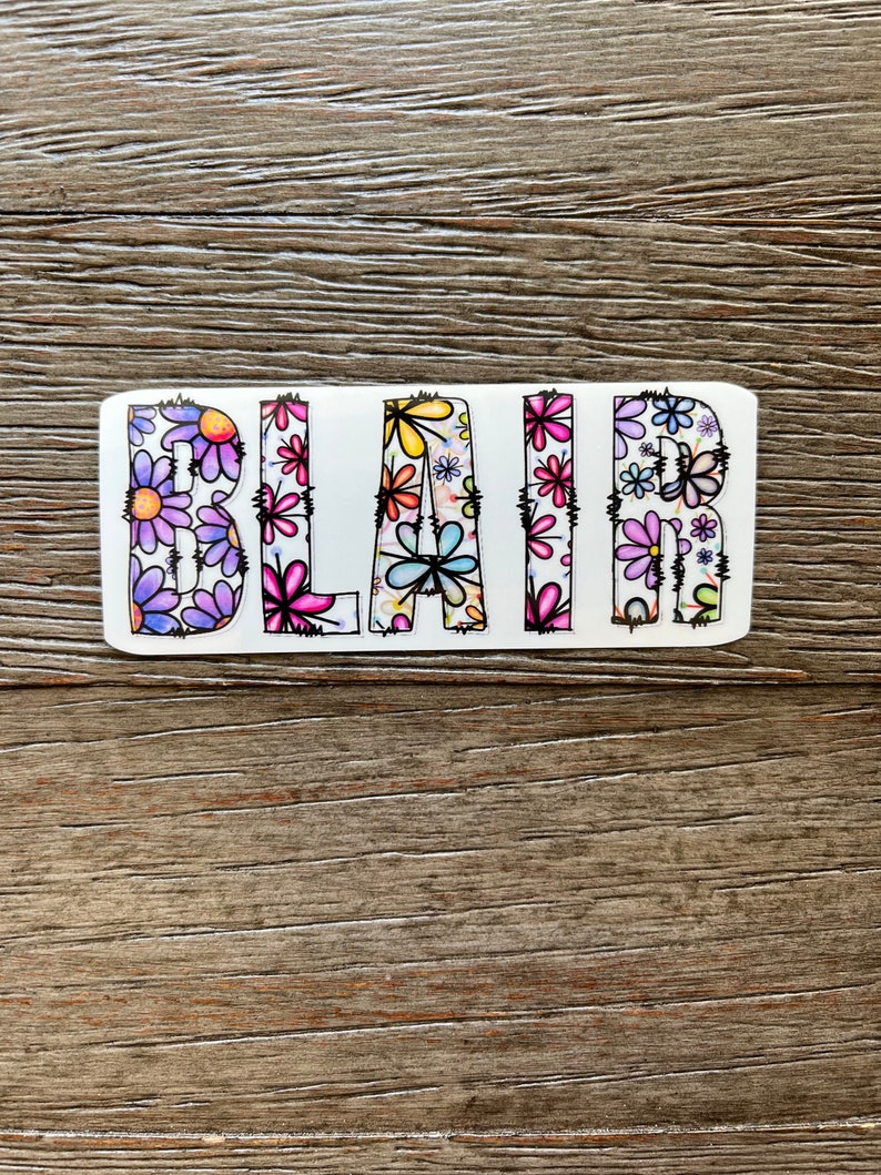 Pode incluir: Um adesivo branco com o nome "BLAIR" escrito em letras pretas delineadas. As letras s&atilde;o preenchidas com um padr&atilde;o floral colorido.