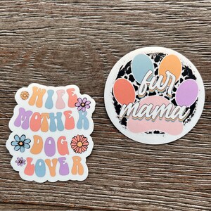 K&ouml;nnte beinhalten: Zwei bunte Aufkleber mit einem Pfotenabdruck-Design. Der erste Aufkleber sagt "Wife Mother Dog Lover" in einer Retro-Schriftart mit Blumen. Der zweite Aufkleber sagt "Fur Mama" mit einem Pfotenabdruck im Hintergrund.