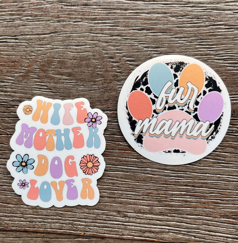 K&ouml;nnte beinhalten: Zwei bunte Aufkleber mit einem Pfotenabdruck-Design. Der erste Aufkleber sagt "Wife Mother Dog Lover" in einer Retro-Schriftart mit Blumen. Der zweite Aufkleber sagt "Fur Mama" mit einem Pfotenabdruck im Hintergrund.