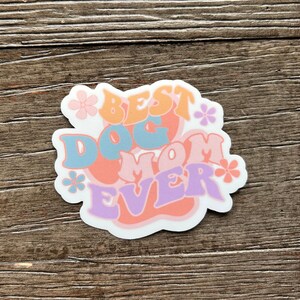 K&ouml;nnte beinhalten: Ein wei&szlig;er Aufkleber mit dem Text "Best Dog Mom Ever" in bunten, stilisierten Buchstaben. Der Text ist von kleinen rosa Blumen umgeben.