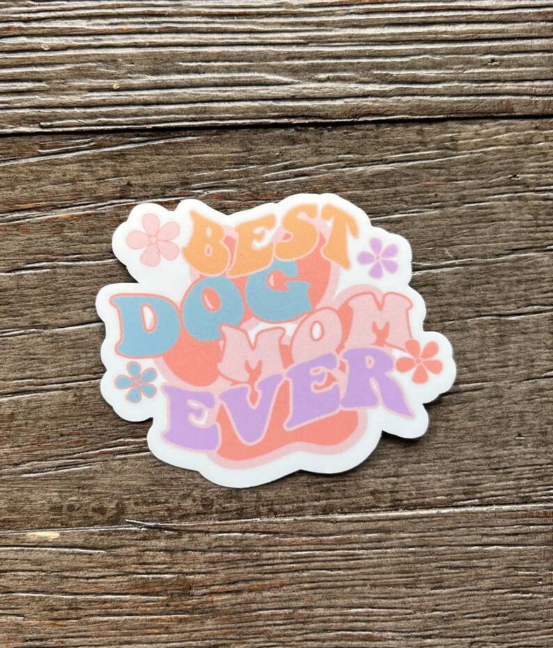 K&ouml;nnte beinhalten: Ein wei&szlig;er Aufkleber mit dem Text "Best Dog Mom Ever" in bunten, stilisierten Buchstaben. Der Text ist von kleinen rosa Blumen umgeben.