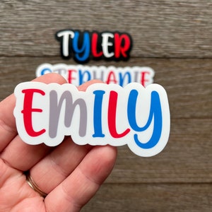 May include: A white sticker with the name "Emily" written in red, gray, and blue letters. The sticker is held in a hand.