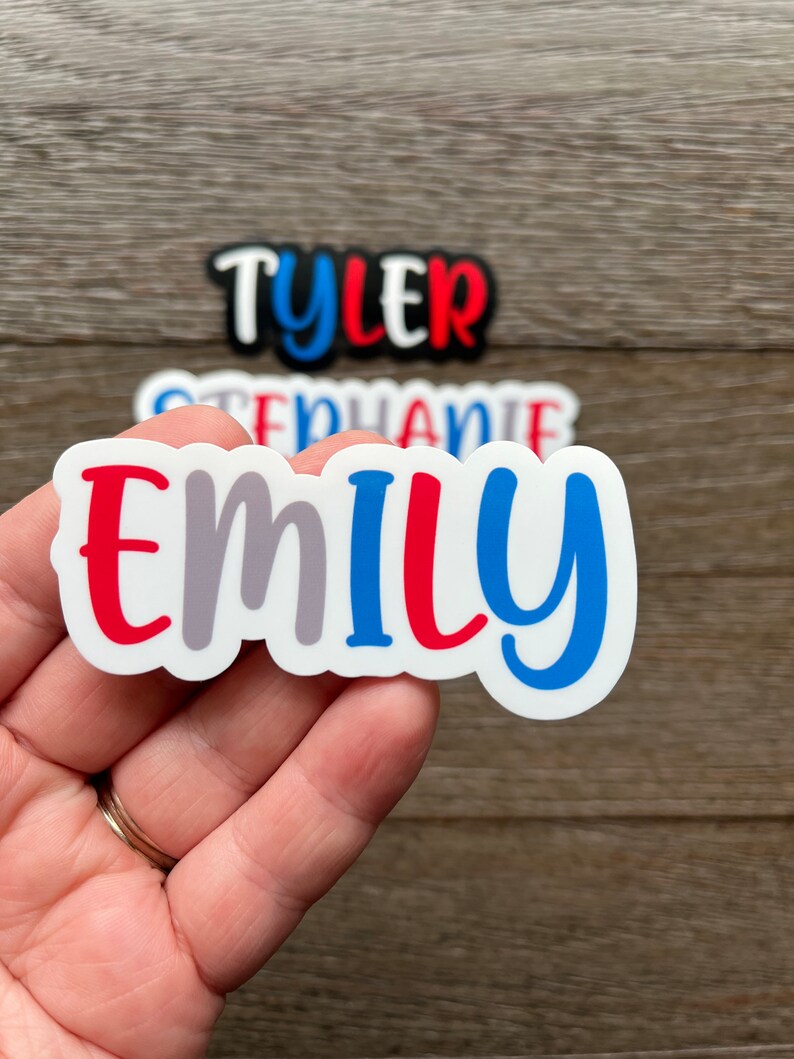 May include: A white sticker with the name "Emily" written in red, gray, and blue letters. The sticker is held in a hand.