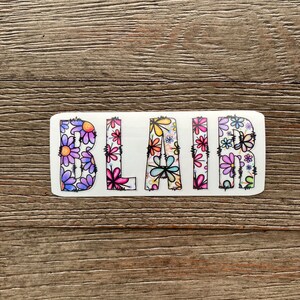 Pode incluir: Um adesivo branco com o nome "BLAIR" escrito em letras pretas delineadas. As letras s&atilde;o preenchidas com um padr&atilde;o floral colorido.