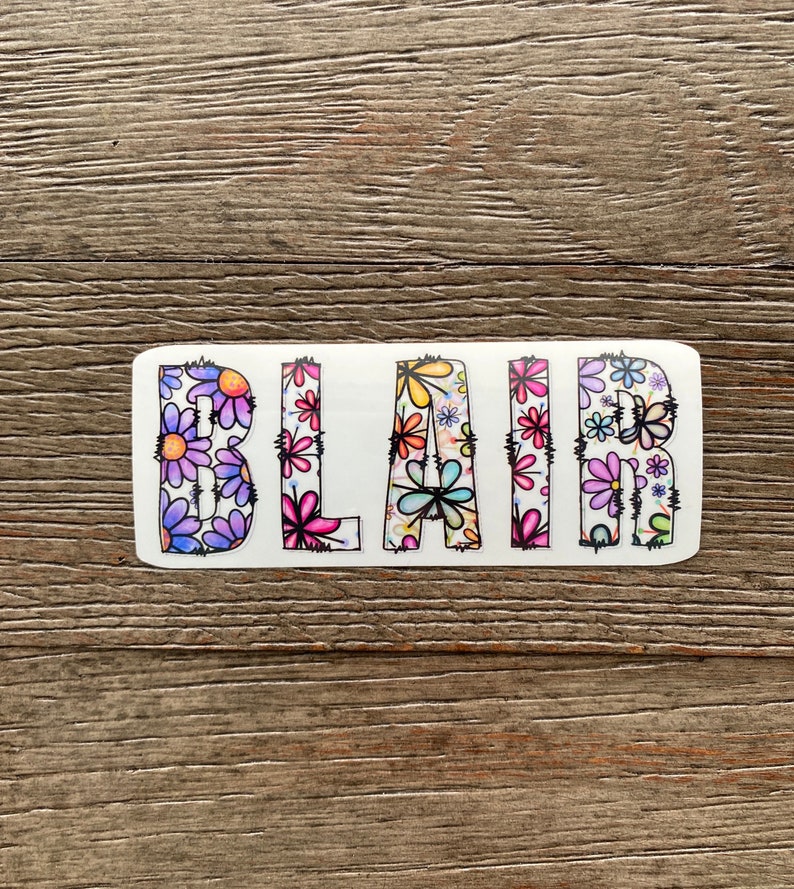 Pode incluir: Um adesivo branco com o nome "BLAIR" escrito em letras pretas delineadas. As letras s&atilde;o preenchidas com um padr&atilde;o floral colorido.
