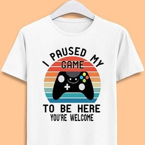 Puede incluir: Camiseta blanca con un gráfico de puesta de sol retro y un mando de videojuego. El texto dice "Pausé mi juego para estar aquí, eres bienvenido".