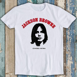 May include: A white short-sleeve t-shirt featuring a black graphic portrait of Jackson Browne centred on the chest. The text JACKSON BROWNE is printed in red above the portrait, and LOS ANGELES, CALIFORNIA is printed in black below the portrait.