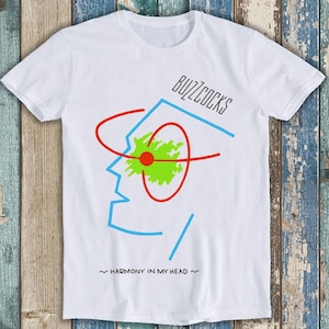 May include: White t-shirt with a graphic design of a stylized face with a red circle and green splatter. The text "Buzzcocks" and "~Harmony in my head~" is printed on the shirt.