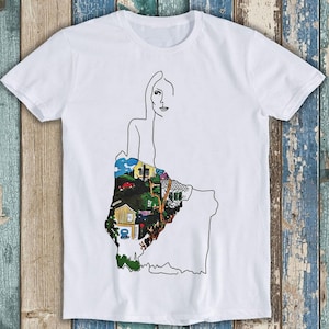 May include: White t-shirt with a line drawing of a person. The person's skirt is filled with a colorful illustration of a town with houses, trees, and cars.