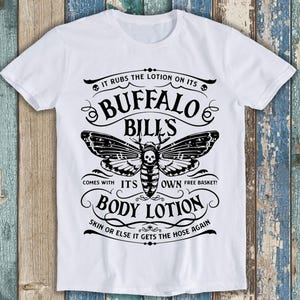 May include: White t-shirt with a black and white graphic design featuring a moth with a skull and crossbones. The text reads "It rubs the lotion on its Buffalo Bills. Comes with its own free basket! Body Lotion. Skin or else it gets the hose again."