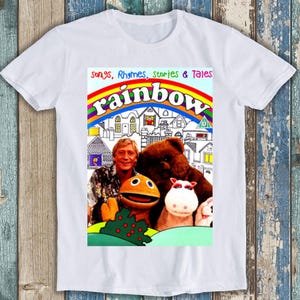 Könnte beinhalten: Weißes T-Shirt mit einer bunten Regenbogengrafik mit dem Text "Songs, Rhymes, Stories & Tales Rainbow". Die Grafik enthält auch ein Cartoon-Bild eines Mannes, eines Bären, einer Puppe und eines Schweins.