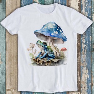 Op de afbeelding: Een wit t-shirt met korte mouwen en een aquarel-stijl afbeelding van een blauwe kikker die onder een grote blauwe paddenstoel met witte stippen zit. Het ontwerp bevat kleine bleke bloemen en een piepkleine rode paddenstoel aan de zijkant.