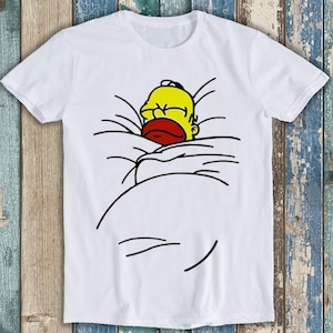 Könnte beinhalten: Ein weißes T-Shirt mit kurzen Ärmeln und einem Grafikdruck einer gelben Zeichentrickfigur mit rotem Mund, die wie unter einer Decke liegend dargestellt ist, wobei schwarze Linien die Falten der Bettwäsche um den Kopf der Figur bilden.