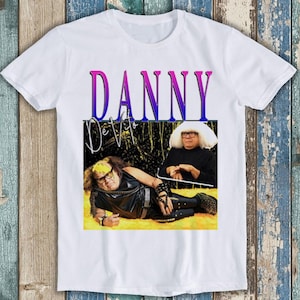 May include: White t-shirt featuring a graphic of Danny DeVito in two different poses. The word "DANNY" is printed in a gradient of pink and purple, with "DeVito" below in a stylized font. The shirt is made of a soft material.