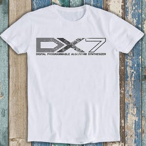 Può includere: Una t-shirt bianca a maniche corte con una stampa grafica nera sul petto. Il design mostra il testo DX7 in un carattere stilizzato a righe orizzontali, con le parole DIGITAL PROGRAMMABLE ALGORITHM SYNTHESIZER stampate al di sotto.