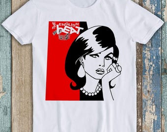 Puede incluir: Una camiseta blanca de manga corta con un estampado gráfico de una persona estilizada con cabello oscuro y un collar de perlas. El diseño incluye un bloque de fondo rojo y negro y el texto THE ENGLISH BEAT en letras blancas.