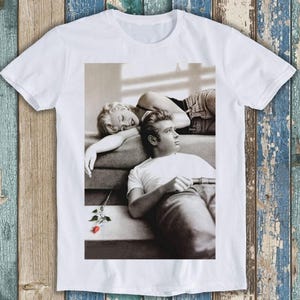 May include: A white short-sleeved t-shirt featuring a black and white photographic print of a person with blonde hair resting their head on the shoulder of another person with dark hair, with a small red rose illustration at the bottom.