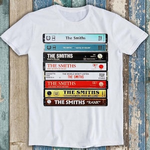 May include: A white short-sleeve t-shirt featuring a graphic print of eight stacked cassette tapes. Each tape label displays The Smiths album titles including Meat Is Murder, The Queen Is Dead, The World Won't Listen, Louder Than Bombs, and Rank.