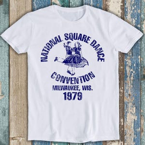 May include: A white short-sleeved t-shirt featuring a distressed blue graphic of a couple square dancing. The text reads National Square Dance Convention Milwaukee, Wis. 1979 in a circular arrangement around the central dancing figures.
