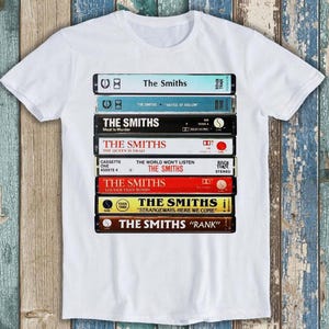 Może przedstawiać: Biała koszulka z nadrukiem stosu kaset magnetofonowych z okładkami albumów The Smiths. Albumy to "Meat is Murder", "The Queen is Dead", "The World Won't Listen", "Louder Than Bombs", "Strangeways, Here We Come" i "Rank".