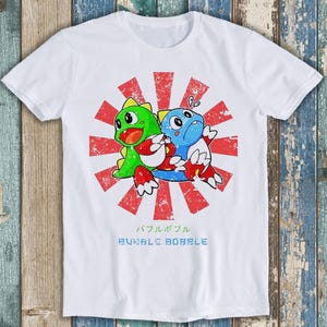 May include: A white short-sleeve t-shirt featuring a graphic of two cartoon dragons, one green and one blue, centered over a distressed red and white sunburst pattern. Below the characters, the text reads "バブルボブル" and "BUBBLE BOBBLE".