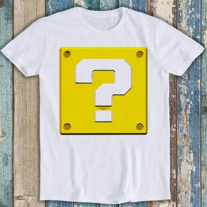 May include: A white short-sleeved t-shirt featuring a large, centred graphic of a yellow square block with a white question mark symbol. The block design includes four small dark circles in each corner, mimicking a metallic or industrial appearance.