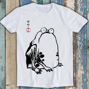 May include: A white short-sleeve t-shirt featuring a black ink-style illustration of a frog with large eyes. To the left of the frog, there is black Chinese calligraphy text positioned above a small red circular stamp with white characters inside.