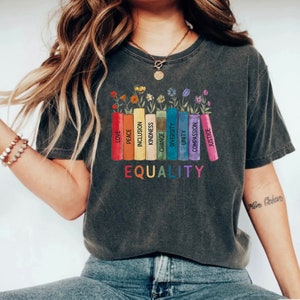 K&ouml;nnte beinhalten: Ein dunkelgraues T-Shirt mit einer Grafik von bunten B&uuml;chern mit W&ouml;rtern wie "Liebe", "Frieden", "Inklusion", "Freundlichkeit", "Ver&auml;nderung", "Vielfalt", "Einheit", "Mitgef&uuml;hl" und "Gerechtigkeit", die auf den Buchr&uuml;cken gedruckt sind. Die W&ouml;rter "GLEICHHEIT" sind unter den B&uuml;chern gedruckt.