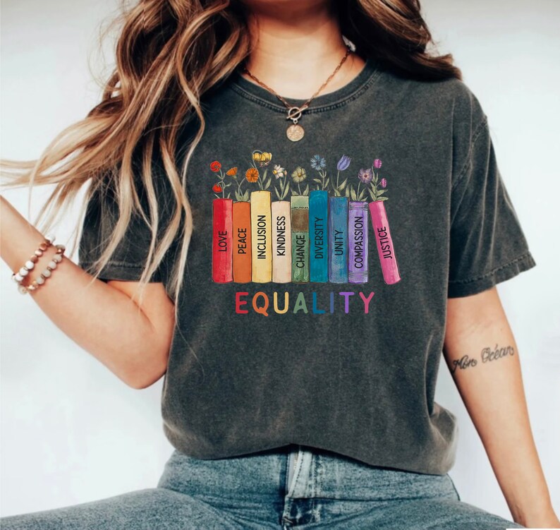 K&ouml;nnte beinhalten: Ein dunkelgraues T-Shirt mit einer Grafik von bunten B&uuml;chern mit W&ouml;rtern wie "Liebe", "Frieden", "Inklusion", "Freundlichkeit", "Ver&auml;nderung", "Vielfalt", "Einheit", "Mitgef&uuml;hl" und "Gerechtigkeit", die auf den Buchr&uuml;cken gedruckt sind. Die W&ouml;rter "GLEICHHEIT" sind unter den B&uuml;chern gedruckt.