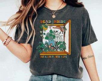 Pode incluir: Uma camiseta cinza com um gráfico de estilo vintage de um esqueleto regando plantas em uma janela. O texto na camiseta diz "Dead Inside But Keeps Plants Alive".