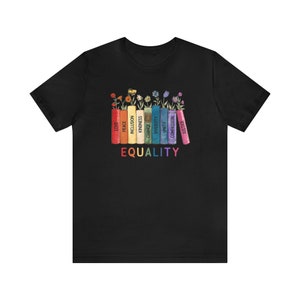 K&ouml;nnte beinhalten: Schwarzes T-Shirt mit einem regenbogenfarbenen Stapel B&uuml;cher mit W&ouml;rtern wie "Liebe", "Frieden", "Inklusion", "Freundlichkeit", "Ver&auml;nderung", "Vielfalt", "Einheit", "Mitgef&uuml;hl" und "Gerechtigkeit" auf den R&uuml;ckseiten. Die W&ouml;rter sind in einem Regenbogenmuster angeordnet. Der Text "GLEICHHEIT" ist unter den B&uuml;chern gedruckt.