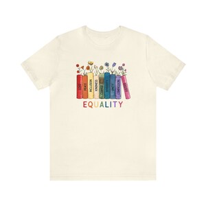 K&ouml;nnte beinhalten: Ein wei&szlig;es T-Shirt mit einem regenbogenfarbenen Design von B&uuml;chern mit W&ouml;rtern wie "Liebe", "Frieden", "Inklusion", "Ver&auml;nderung", "Freundlichkeit", "Vielfalt", "Einheit", "Mitgef&uuml;hl" und "Gerechtigkeit" auf den R&uuml;ckseiten. Die W&ouml;rter sind in einem Regenbogenmuster angeordnet und die B&uuml;cher sind mit Blumen verziert. Die W&ouml;rter "GLEICHHEIT" sind unter den B&uuml;chern gedruckt.
