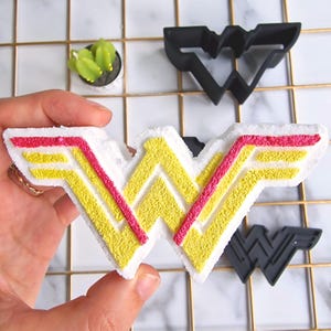 Könnte beinhalten: Ein weißer Badebombe in Form des Wonder Woman-Logos. Das Logo ist rot und gelb umrandet.