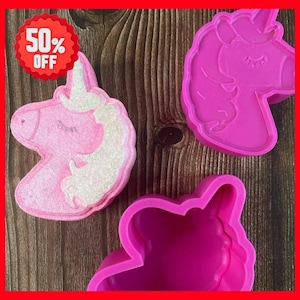 Op de afbeelding: Drie items met een eenhoornthema worden getoond: een roze badbruisbal en twee roze plastic mallen. De badbruisbal heeft een witte manen en hoorn. Een rode cirkel met "50% OFF" staat in de linkerbovenhoek.