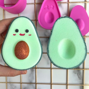 Könnte beinhalten: Zwei grüne Avocado-Badebomben mit dunkelgrünem Umriss. Eine Avocado hat eine braune Mitte und ein lächelndes Gesicht mit rosa Wangen. Die andere Avocado hat eine braune Mitte.