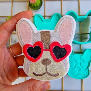 May include: A French bulldog-shaped cookie, decorated with heart-shaped sunglasses. The cookie features a white, tan, and brown colour palette, with red and black details. A teal cookie cutter is in the background.