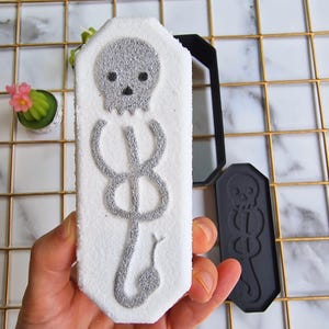 Può includere: Un oggetto rettangolare bianco con un disegno di teschio e serpente argentato. Accanto c'è uno stampo rettangolare nero con lo stesso disegno. Il disegno è un teschio sopra un serpente, un simbolo delle Arti Oscure.