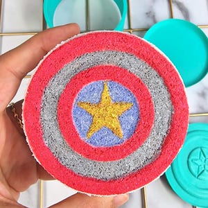Könnte beinhalten: Eine rot-weiß-blaue Badebombe in Form von Captain Americas Schild. Der Schild hat einen fünfzackigen goldenen Stern in der Mitte.