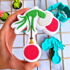 May include: A white bath bomb shaped like a green character holding a red ornament. The character has a textured green body, a white and red hat, and a gold-colored string detail. The bath bomb is held against a background of teal cookie cutters.