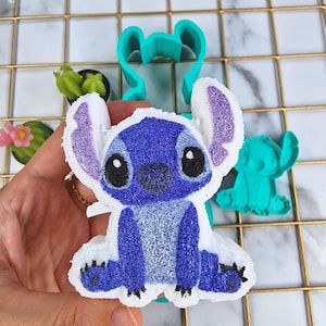 Puede incluir: Bomba de baño azul y blanca con forma de Stitch de Lilo & Stitch. La bomba de baño está sobre un molde de plástico verde de Stitch.
