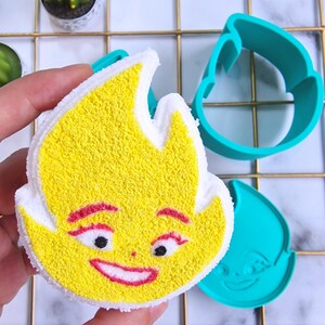 May include: A yellow bath bomb shaped like a flame with a smiling face. The bath bomb is being held in a hand. There are two blue plastic molds in the background, one shaped like a flame and the other shaped like a troll.