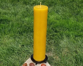 May include: A large, yellow beeswax candle sits on a black metal base atop a gray rock. Five small, reddish-brown stones are arranged around the base of the candle.
